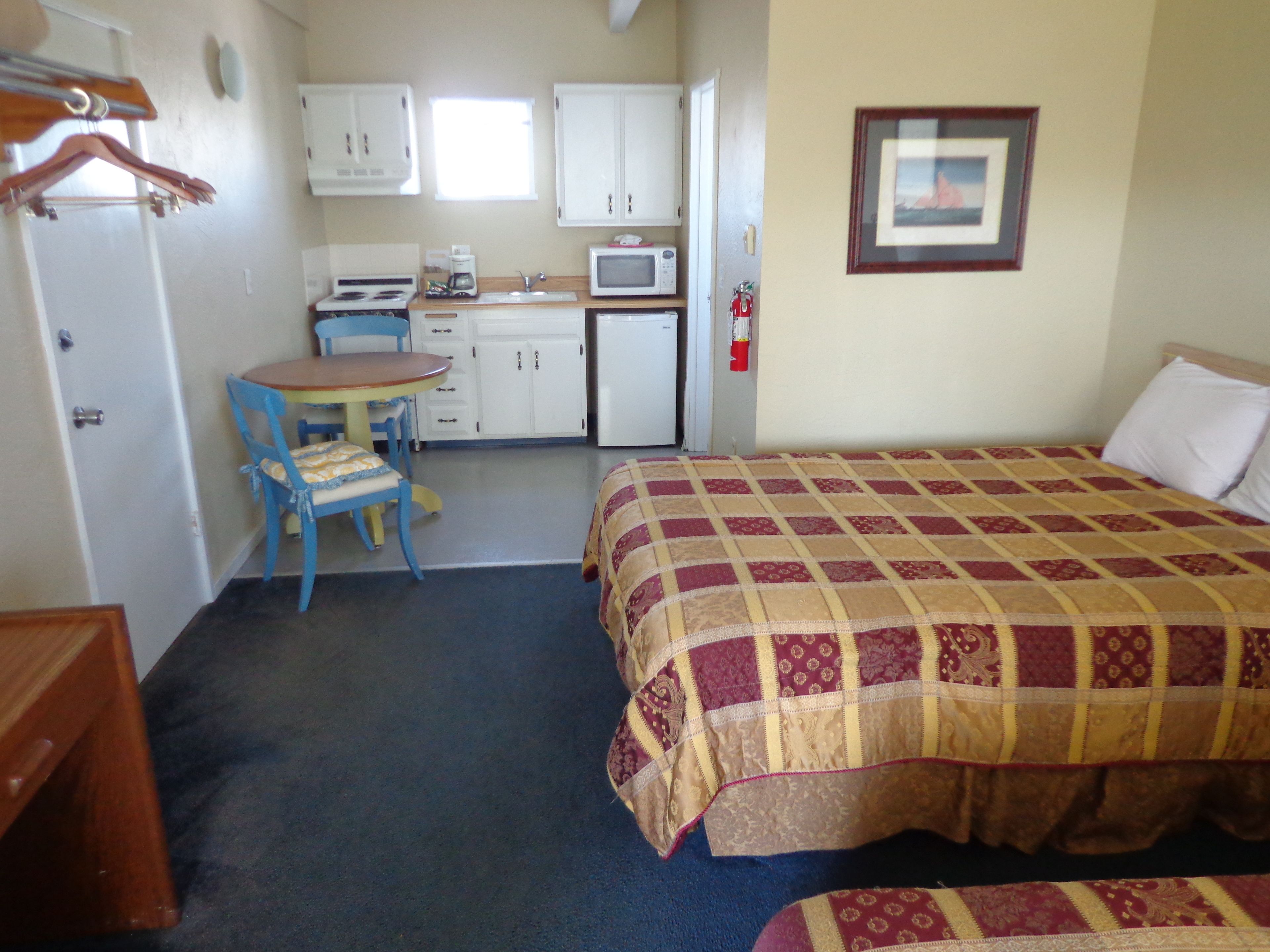 Une pièce de taille modeste comprenant un lit, une petite kitchenette, un coin repas avec une table et des chaises, et une porte avec un porte-manteau.