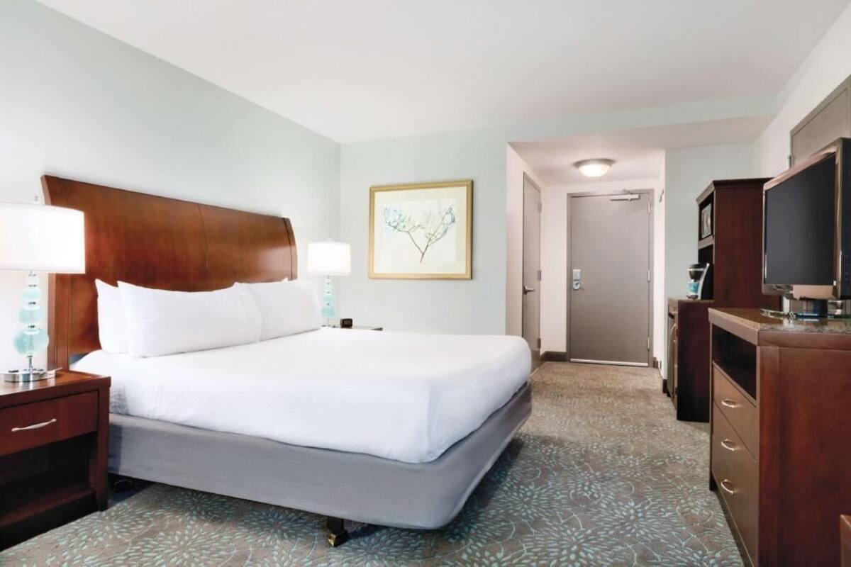 A hotel room with a large bed, bedside lamps, a framed artwork on the wall, a door, and a minibar.
