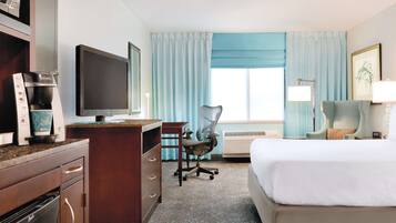 Hotelkamer met bed, bureau, stoel, kitchenette en een groot raam met gordijnen.