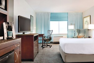 Habitación de hotel con cama, escritorio, silla, kitchenette y un ventanal con cortinas.