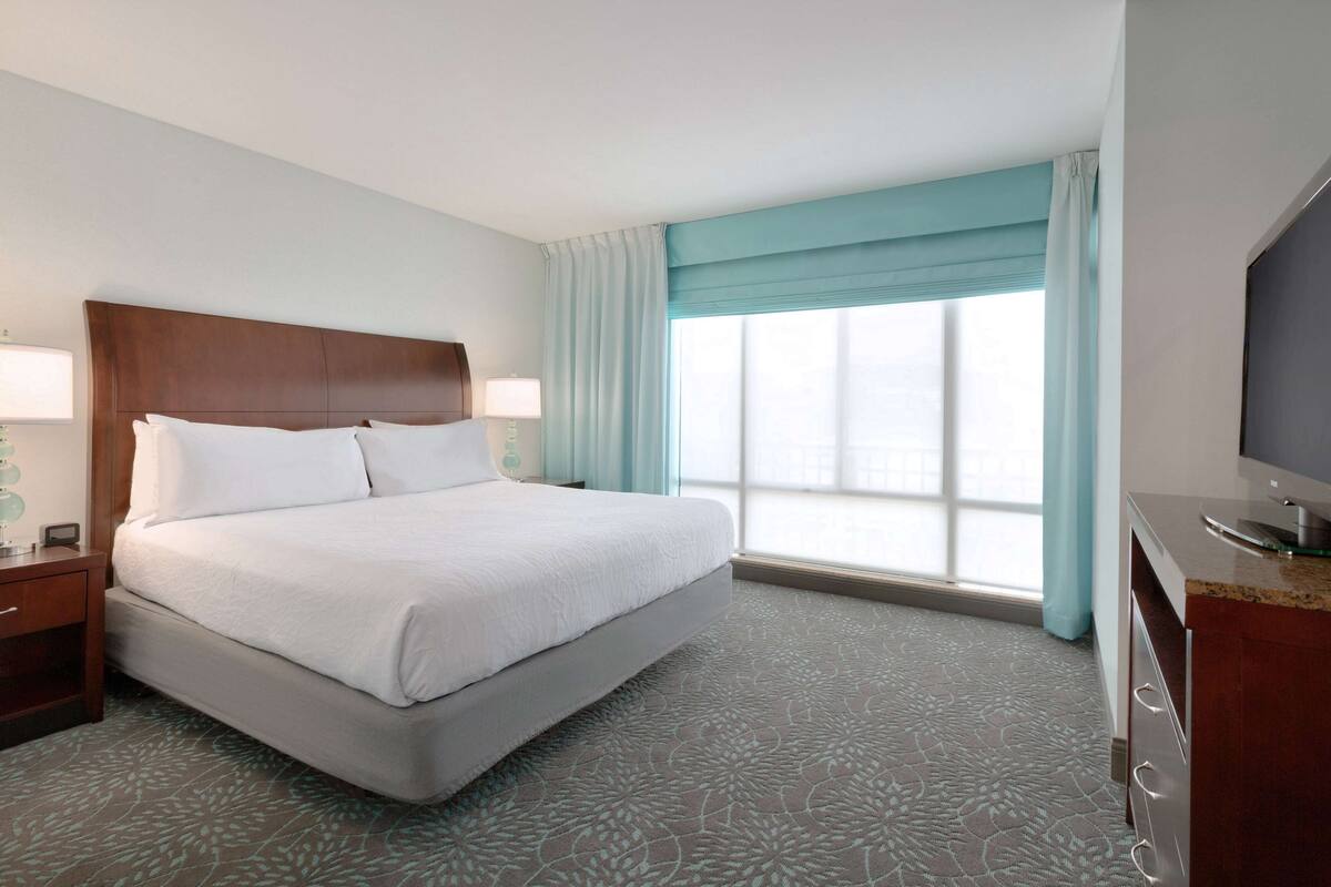 A hotel room with a large bed, a wooden headboard, a nightstand with a lamp, a television, and a window with curtains.