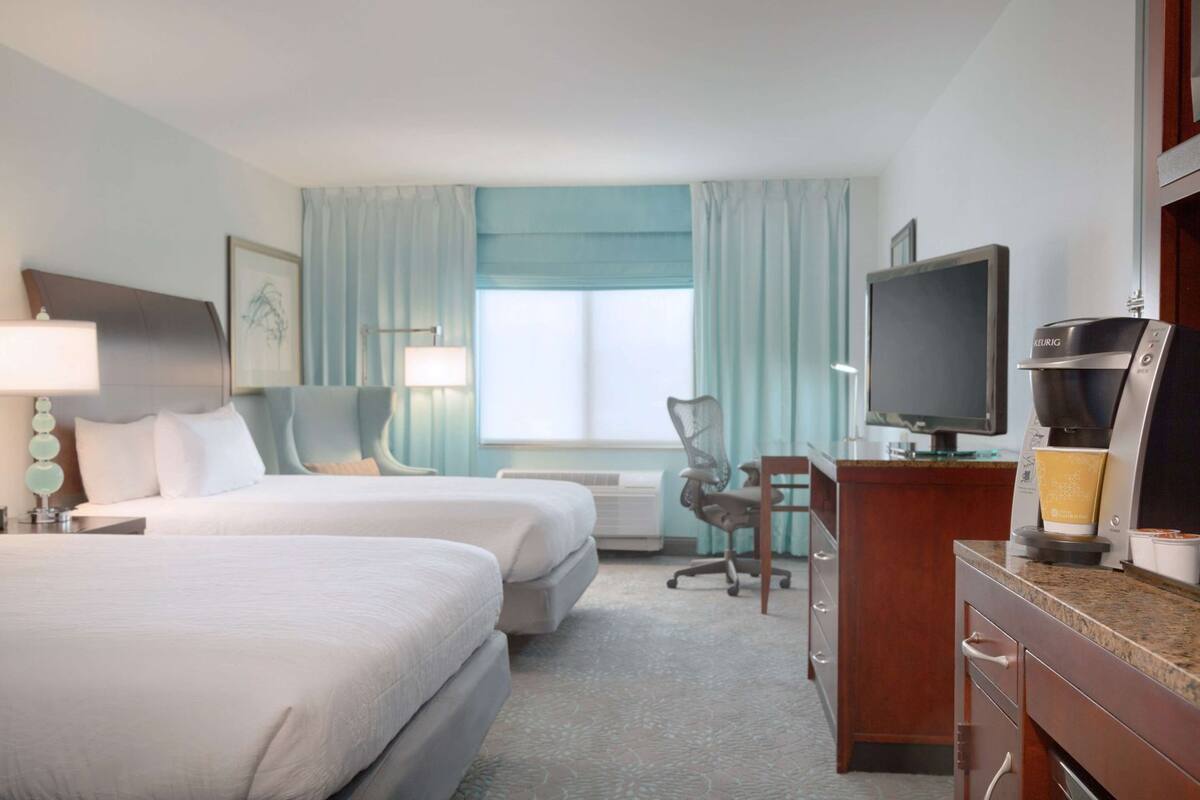 A hotel room with two beds, a desk with a coffee maker, a TV, and a window with curtains.