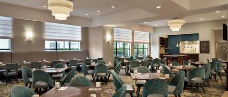 A spacious hotel dining area with multiple tables and chairs, large windows, and a central bar area.