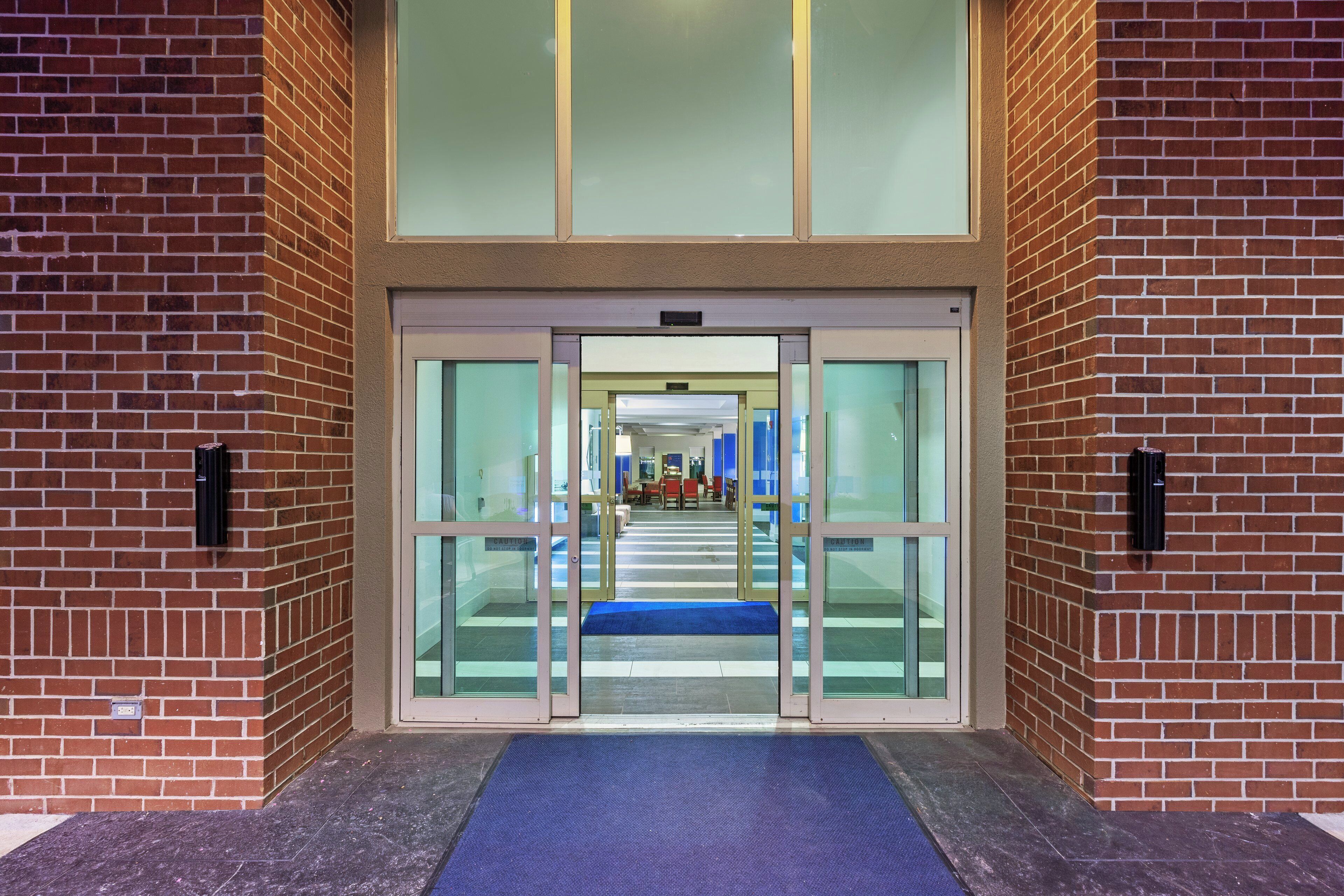 A building entrance with glass doors, a blue mat, and a brick wall.