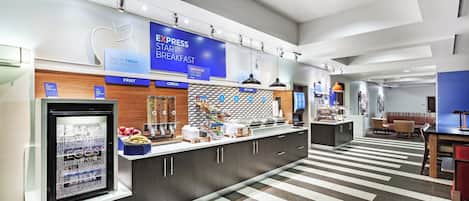 Un área de desayuno con un mostrador de autoservicio, vitrina refrigerada y diversas opciones de alimentos.