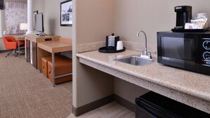 Habitación de hotel compacta con kitchenette, que incluye microondas, cafetera y lavabo. Cuenta con una pequeña zona de estar con una silla y una mesa, y un ventanal con vistas.