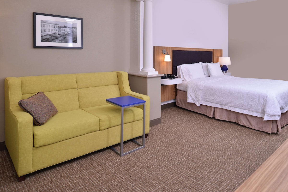 Hotellihuoneessa on keltainen sohva, pieni sivupöytä, valkoisilla vuodevaatteilla varustettu sänky, yöpöytä lampun kanssa ja seinällä oleva kehystetty kuva.