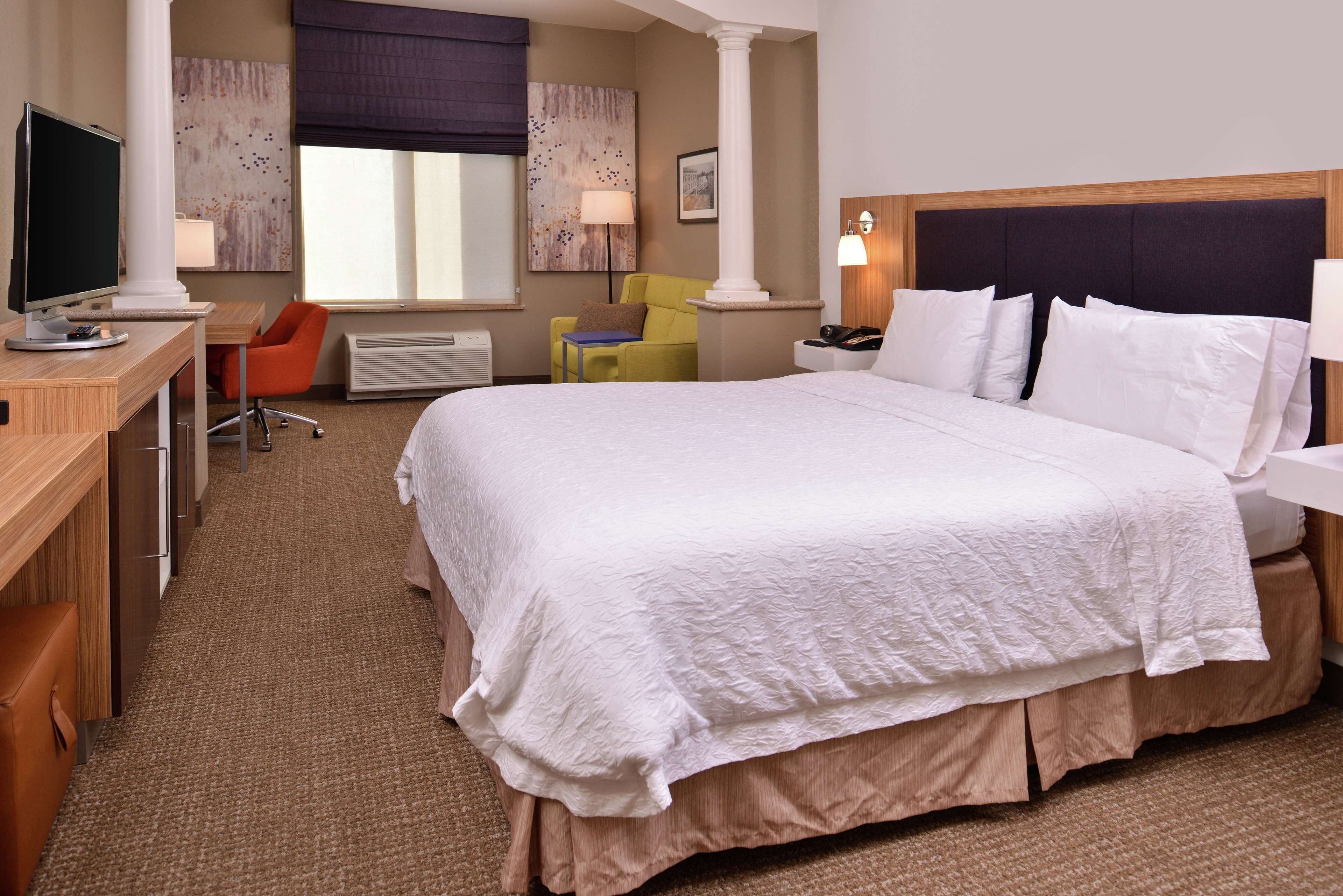 Ein Hotelzimmer mit einem großen Bett, einem Schreibtisch mit Computer, einem Stuhl, einer Lampe und einem Fenster mit Jalousien.