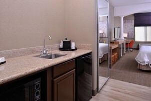 Habitación de hotel con cocina americana que incluye un lavamanos, cafetera y lavavajillas. Cuenta con un escritorio y silla, cama y ventana con vistas.