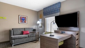 A modern hotel room with a grey sofa, a wooden desk, a flat-screen TV, and a framed picture on the wall. - Hampton Inn & Suites Louisville East (Louisville)