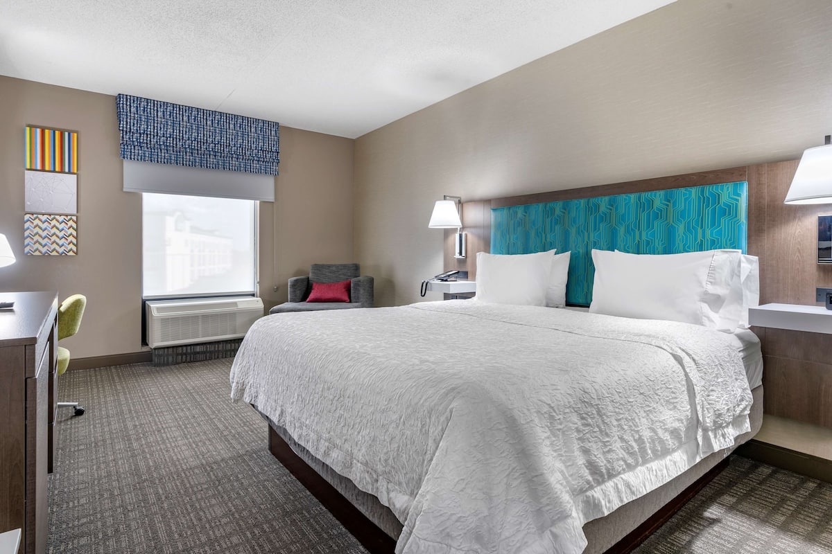 A hotel room with a large bed, a desk with a chair, a window with blinds, and a wall-mounted lamp.