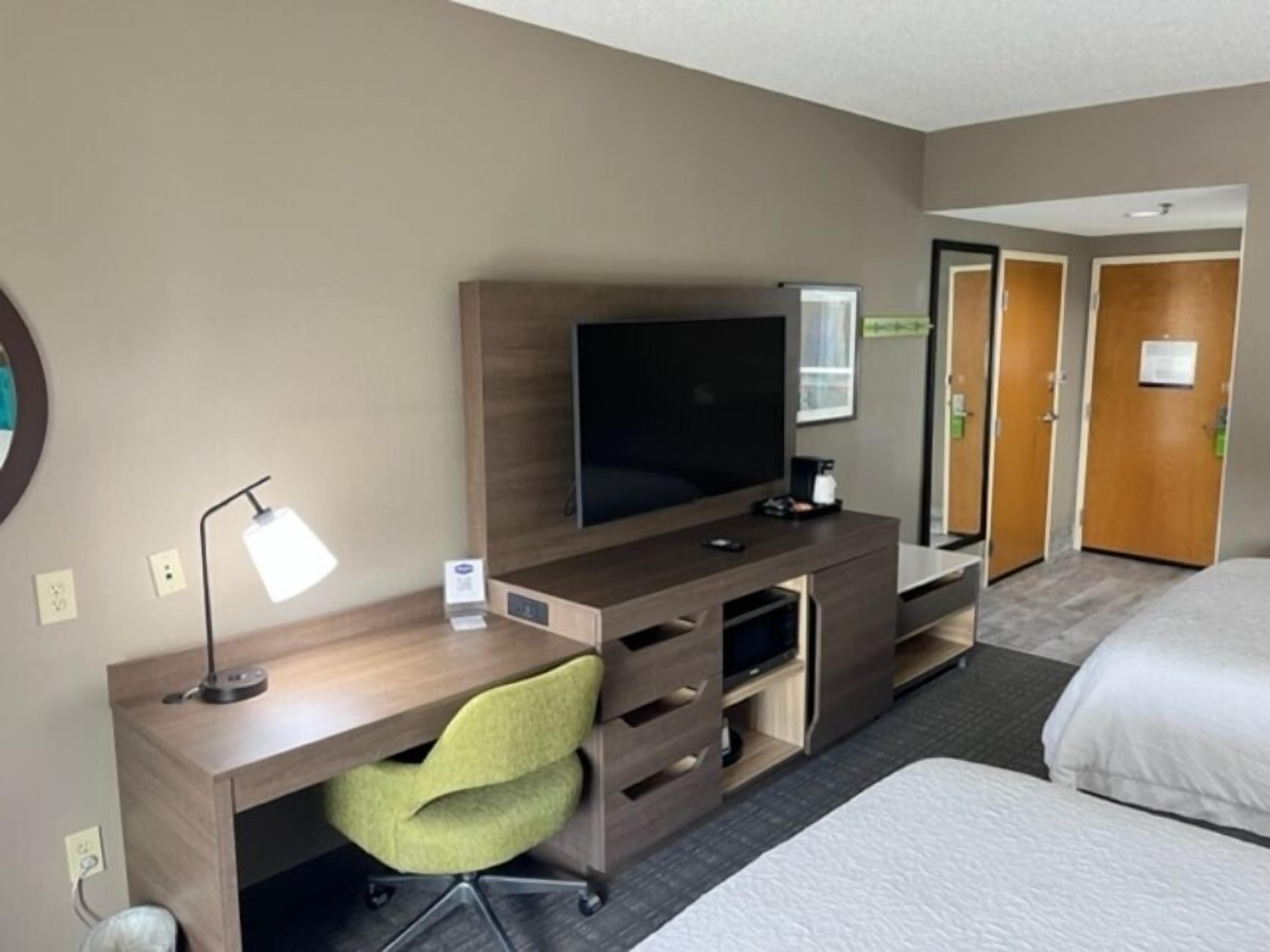 A hotel room with a wooden desk, a flat-screen TV, a green office chair, a bed, and a door leading to a bathroom.