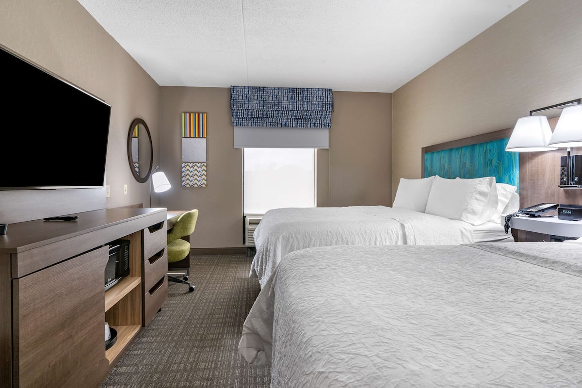 A hotel room with two beds, a desk with a flat-screen TV, a lamp, a chair, and a window with a view.