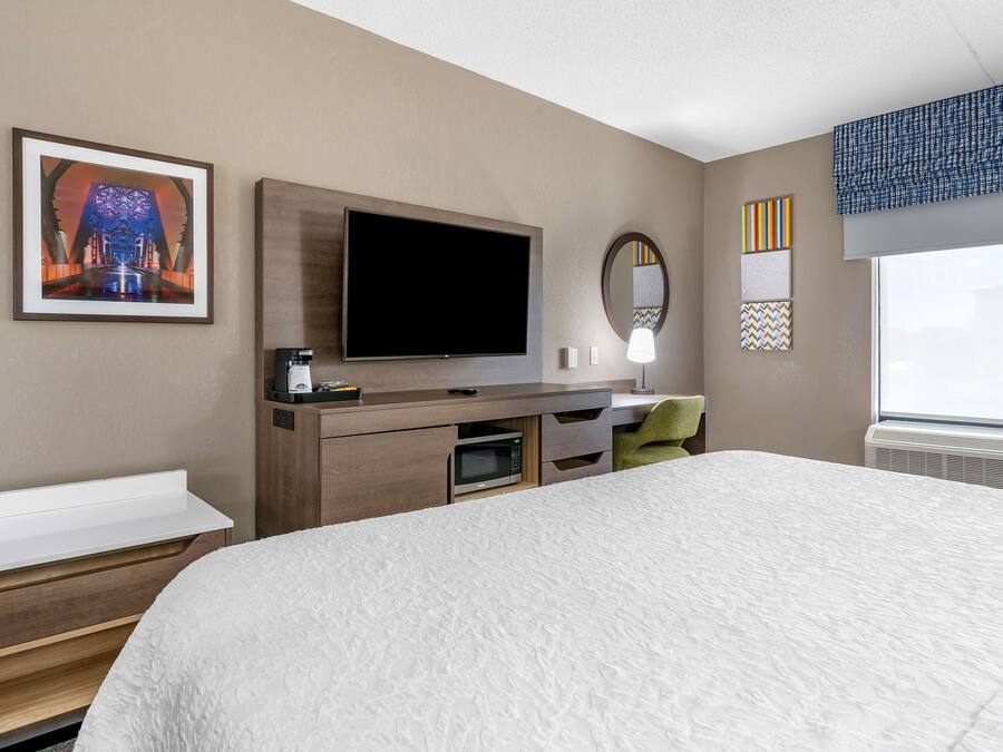 A hotel room with a large bed, a flat-screen TV, a microwave, a small desk, a chair, and a window with blinds.