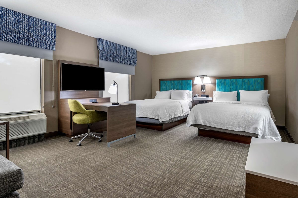 A hotel room with two beds, a desk, a chair, a TV, and a window with blinds.
