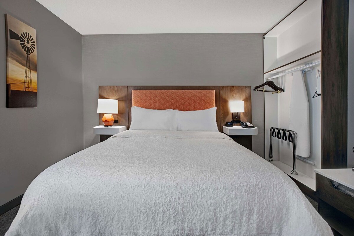 A hotel room with a large bed, two bedside tables with lamps, a headboard with a textured panel, a coat rack, and a window with curtains.