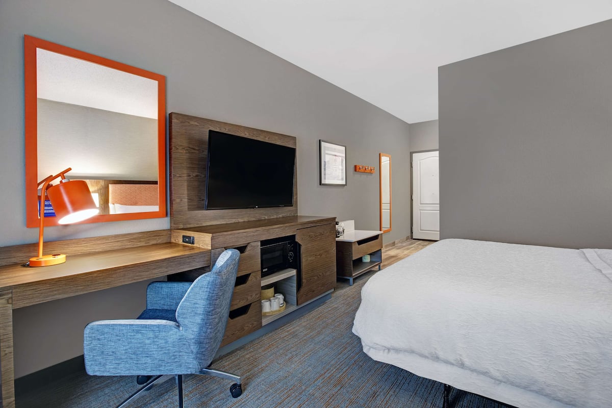 A hotel room with a bed, a desk with a TV, a chair, and a large mirror.