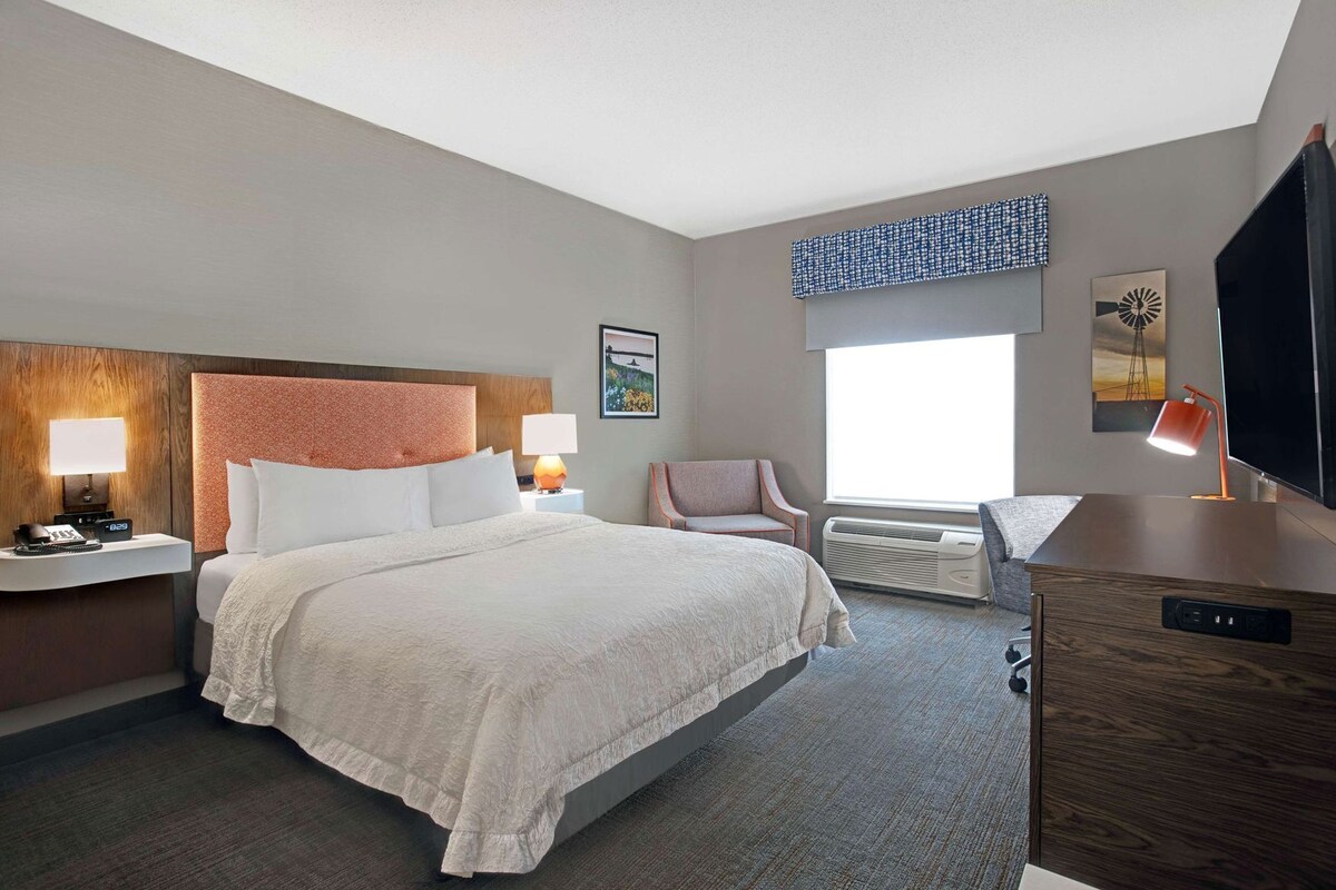 A hotel room with a bed, bedside lamps, a chair, a desk, a television, and a window.