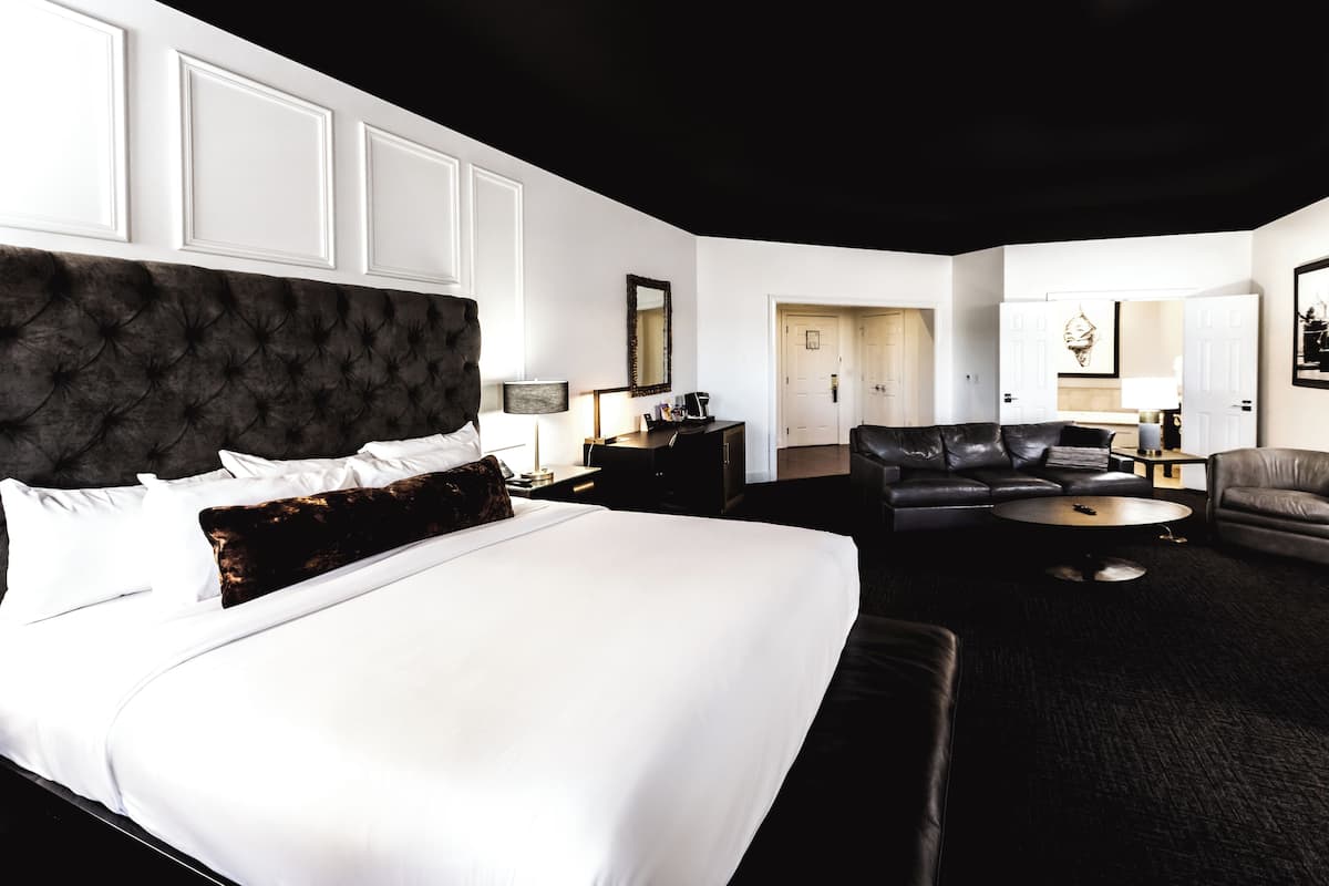 A modern hotel room with a large bed, a dark upholstered headboard, a black leather sofa, and a round coffee table.