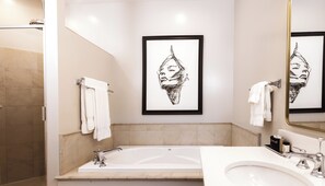 Un baño moderno con bañera, lavamanos y un espejo grande. Hay toallas colgadas en la pared y una obra de arte enmarcada sobre la bañera.