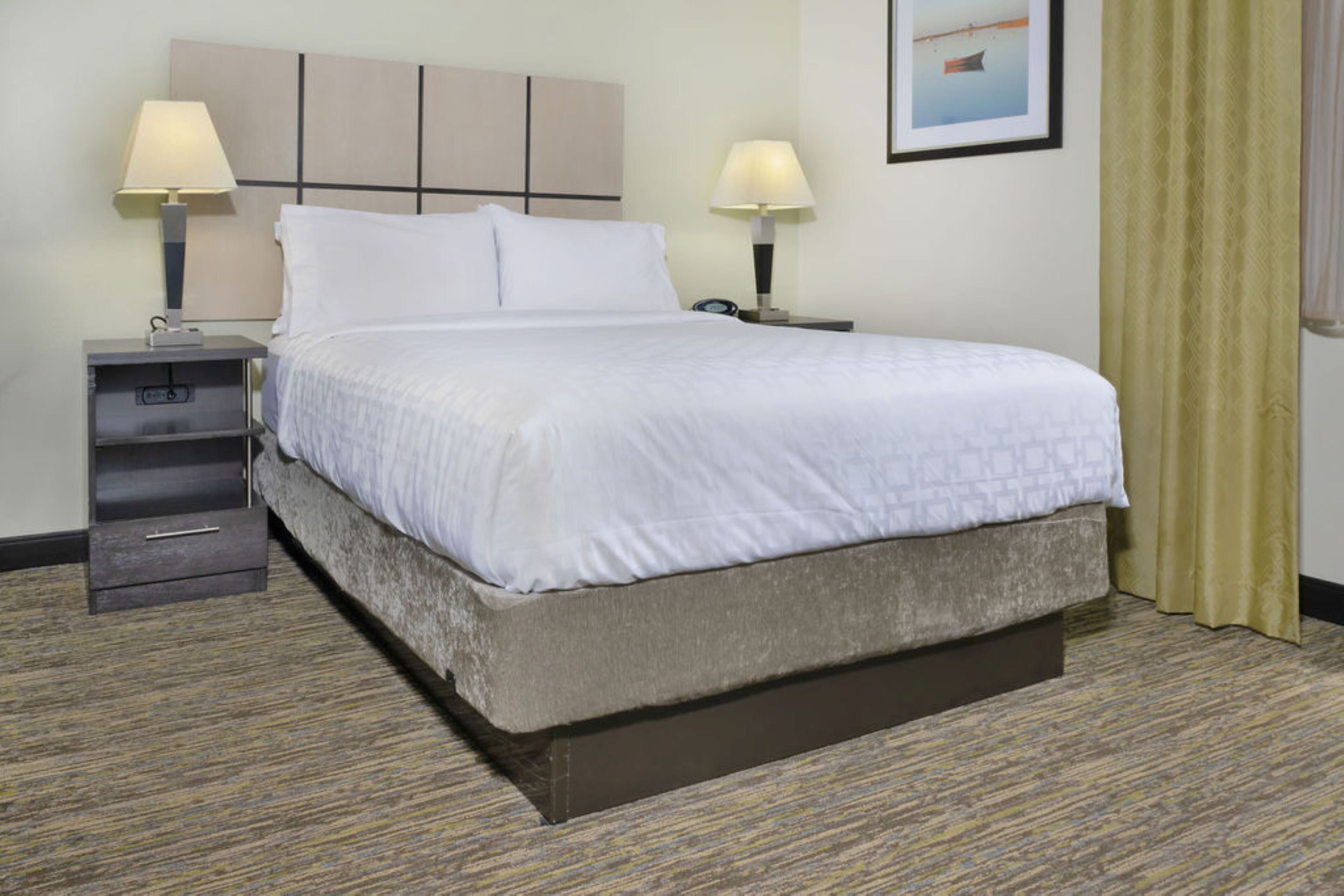 A hotel room with a bed, two bedside tables with lamps, a small drawer, a framed picture on the wall, and a patterned carpet.