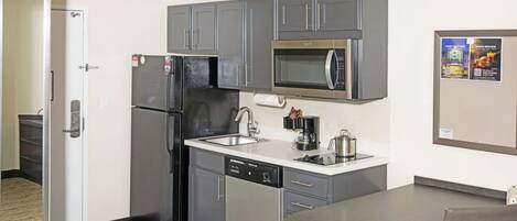 A compact kitchen with grey cabinets, a stainless steel refrigerator, a microwave, a dishwasher, and a small dining table with a chair.