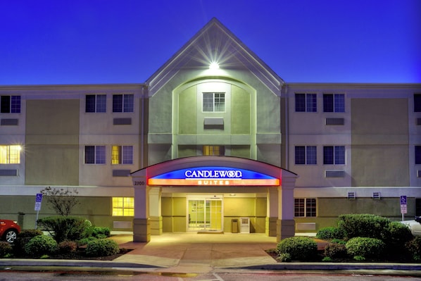A hotel with a prominent entrance, a sign reading "Candlewood Suites," and a parking lot with a red car.