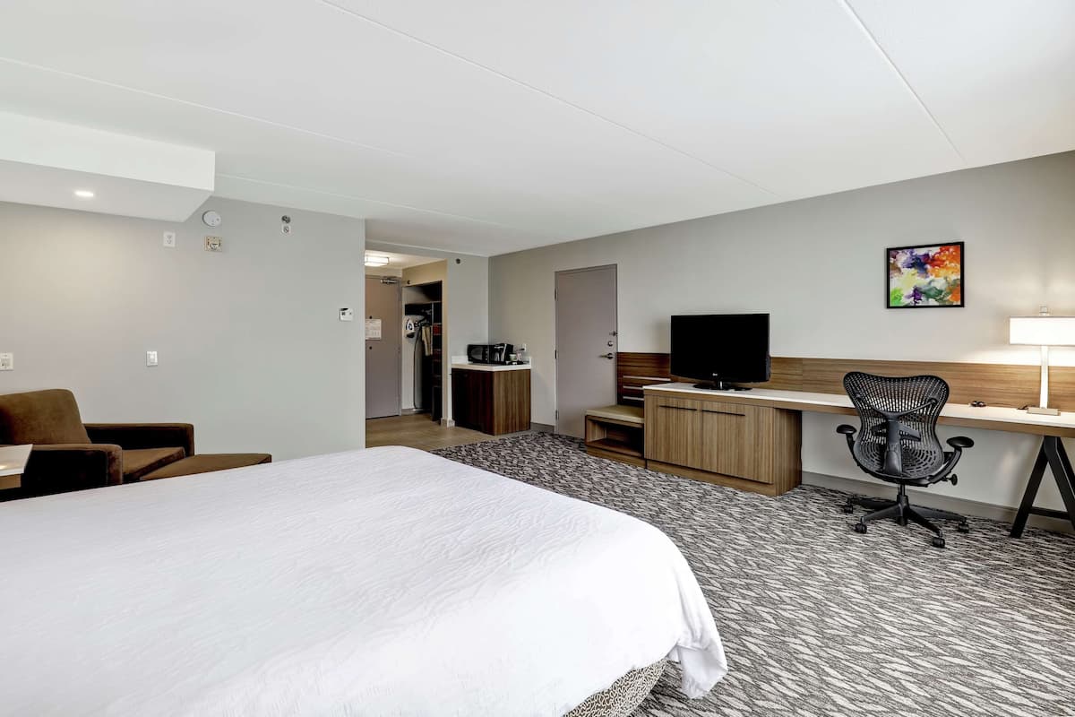 A hotel room with a large bed, a desk with a computer, a chair, a TV, and a small kitchenette.