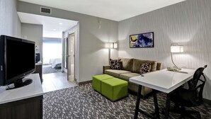 Hotellihuone, jossa on litteänäyttöinen televisio, värikkäillä koristetyynyillä varustettu sohva, pieni pöytä lampun kanssa ja kuviollinen matto.