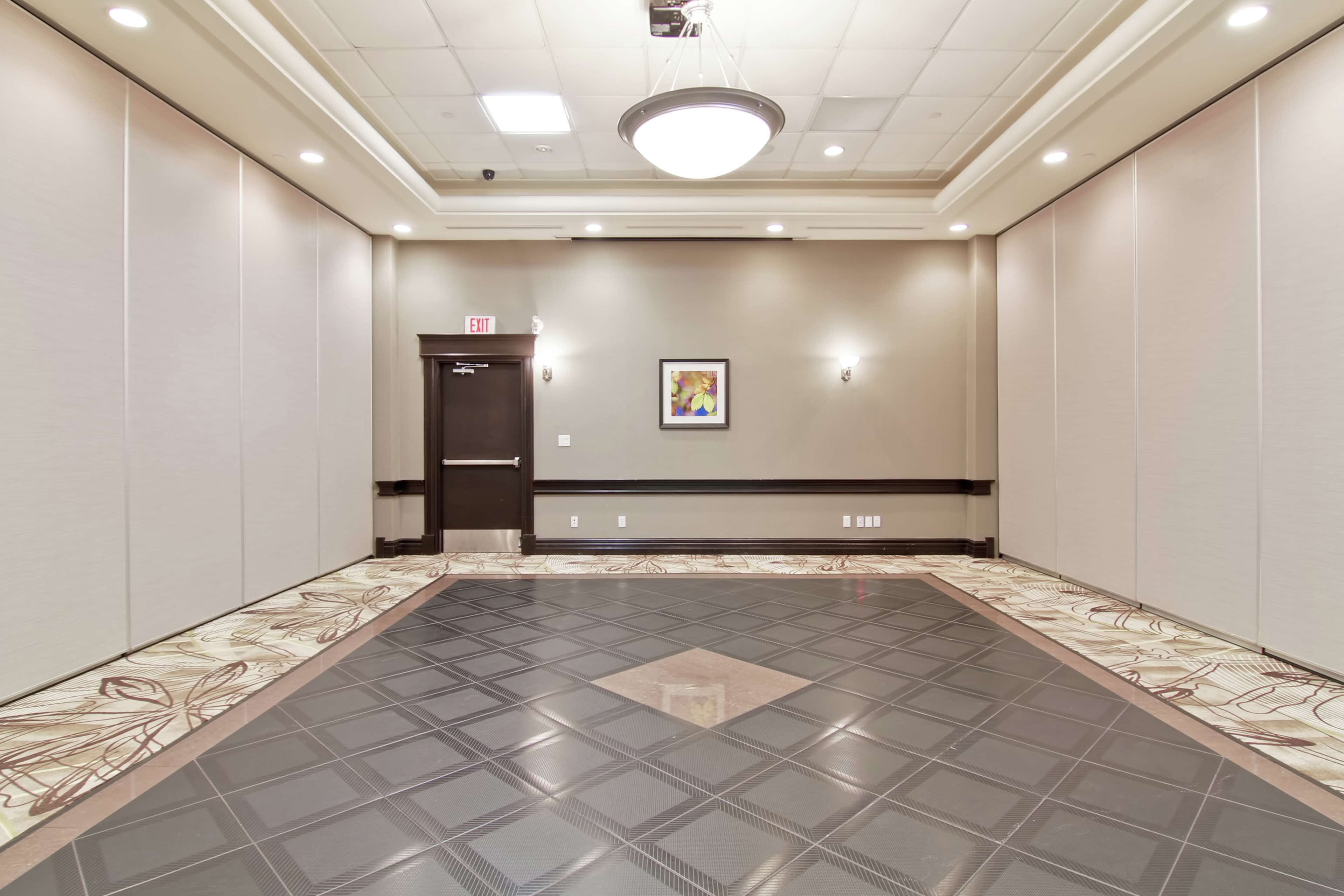 A spacious, well-lit hallway with a patterned floor, a door with an exit sign, and a framed picture on the wall.