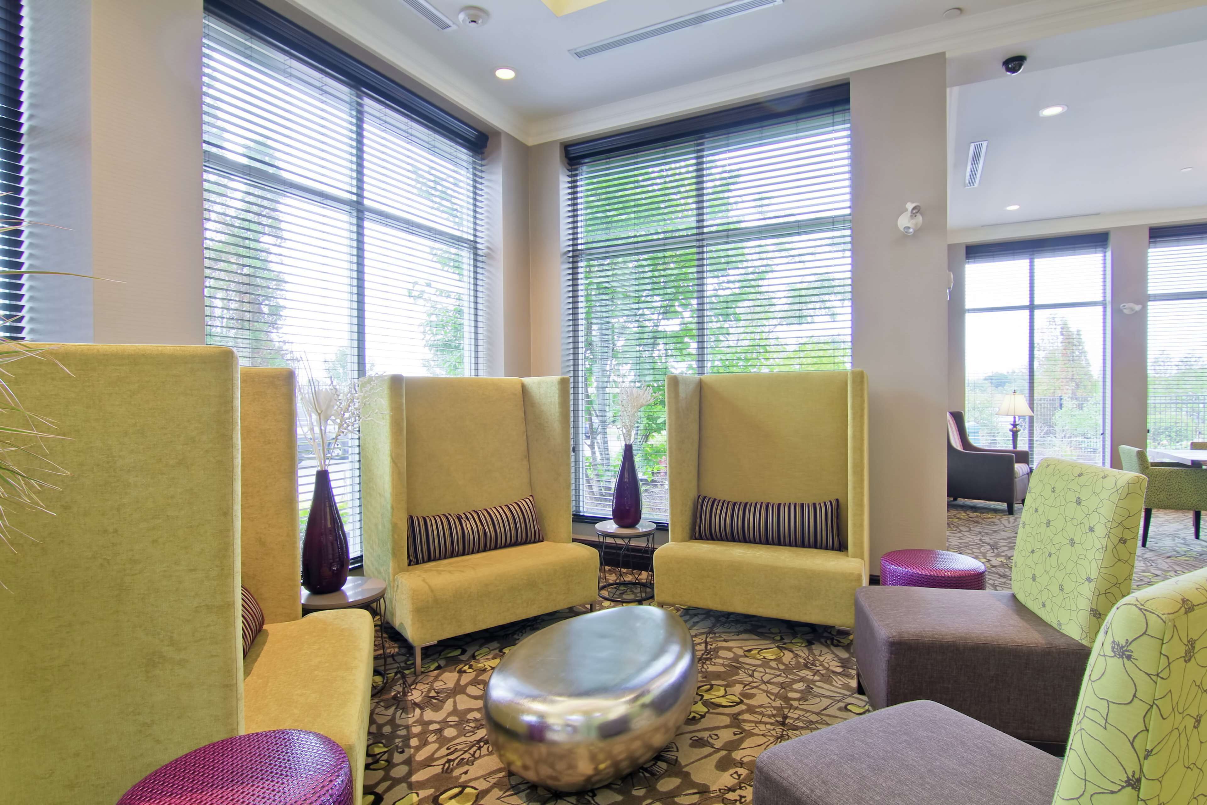A hotel lobby with yellow armchairs, a patterned carpet, and large windows with blinds.