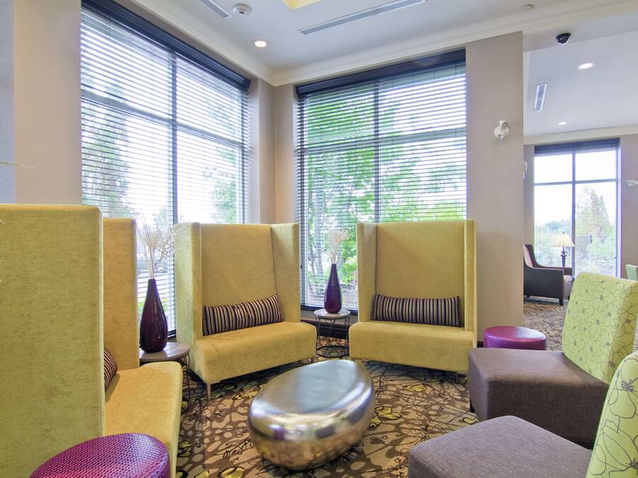Um saguão de hotel com poltronas amarelas, um tapete estampado e janelas grandes com persianas.