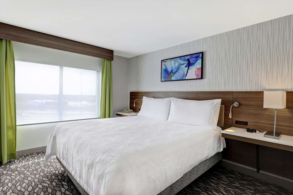 A hotel room with a large bed, a nightstand, a lamp, a painting on the wall, and a view of the airport through the window.