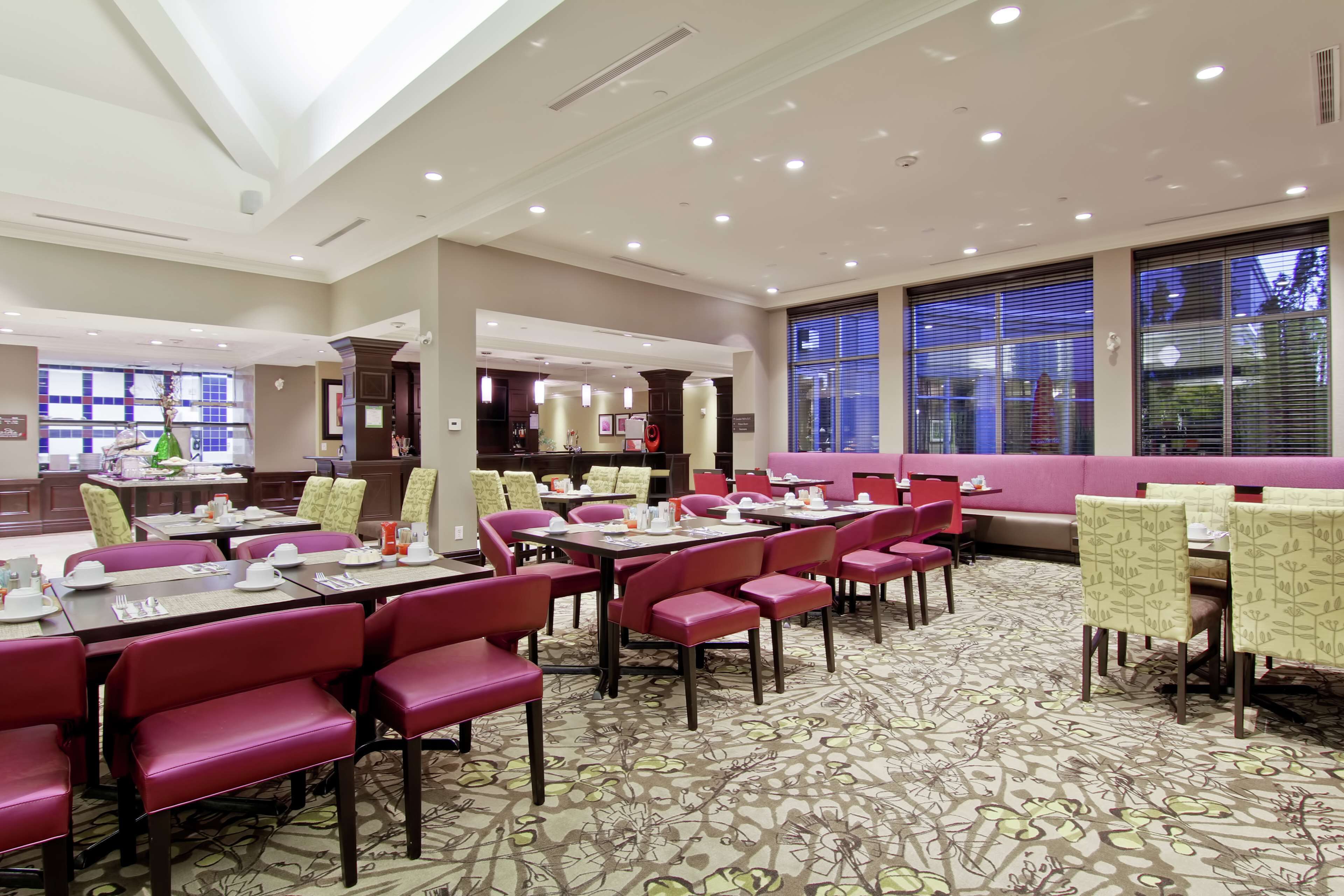 A hotel dining area with tables and chairs, a reception desk, and a large window with blinds.