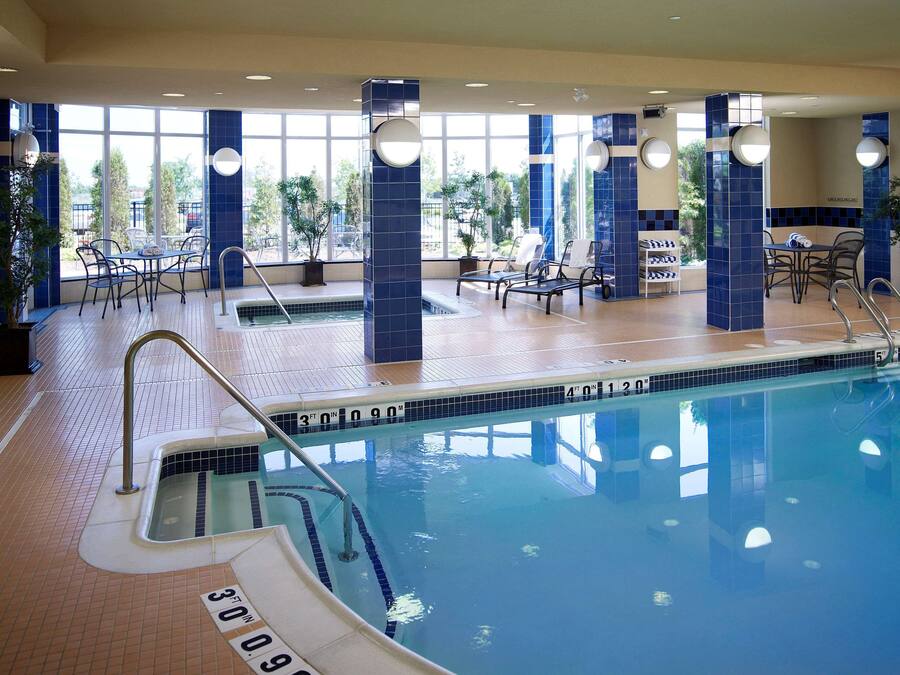 Una piscina interior con agua cristalina, paredes de azulejos y una zona soleada con sillas y mesas.