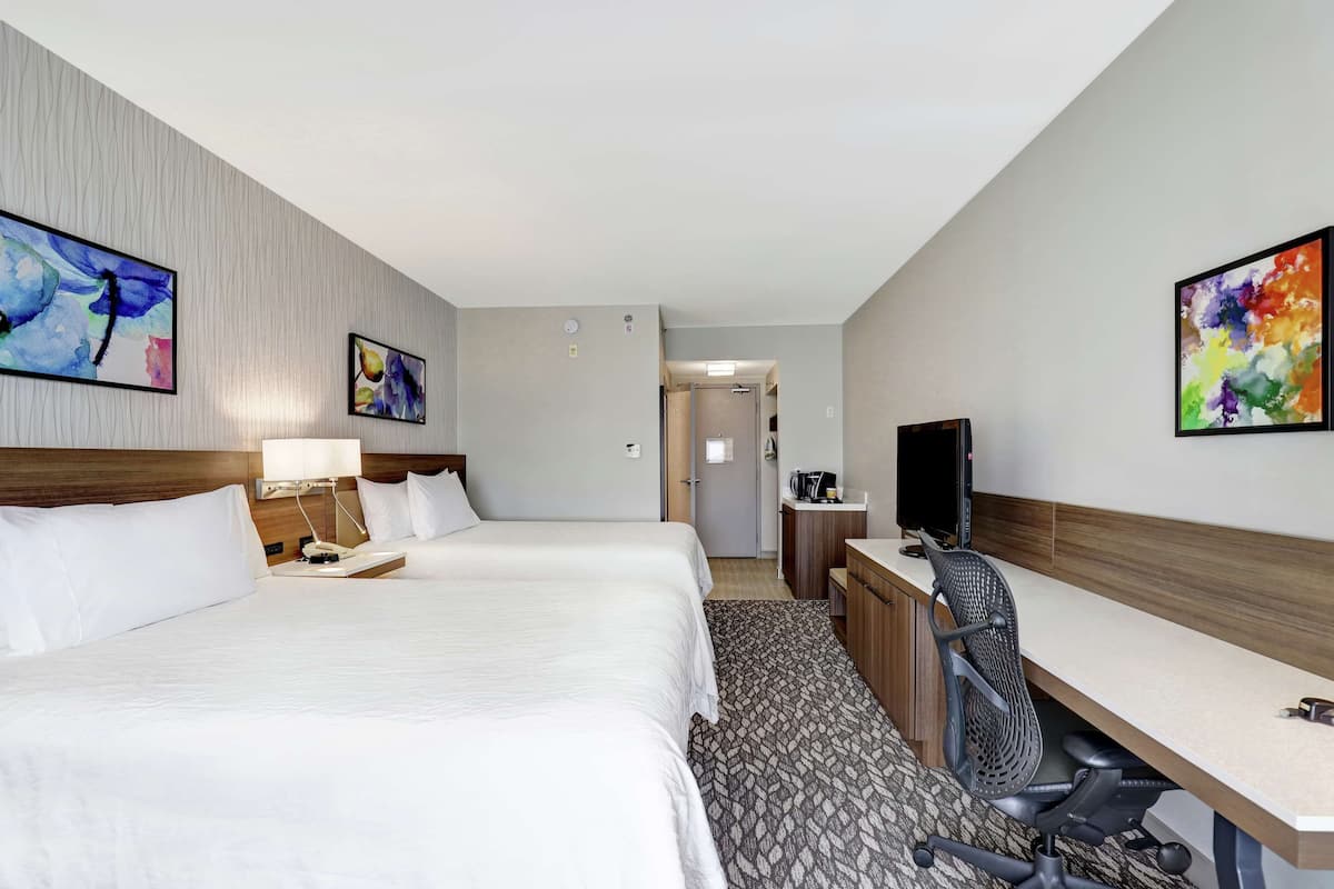 A hotel room with two beds, a desk, a chair, a TV, and artwork on the walls.