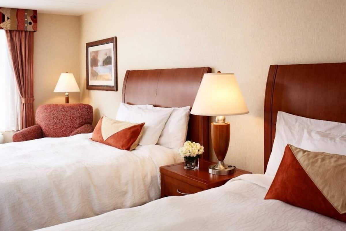 A hotel room with two beds, a nightstand with a lamp, a chair, and a framed picture on the wall.