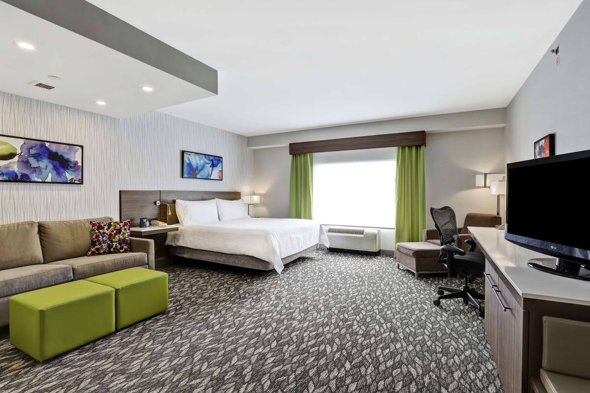 A hotel room with a bed, a sofa, a desk with a TV, and a window with curtains.