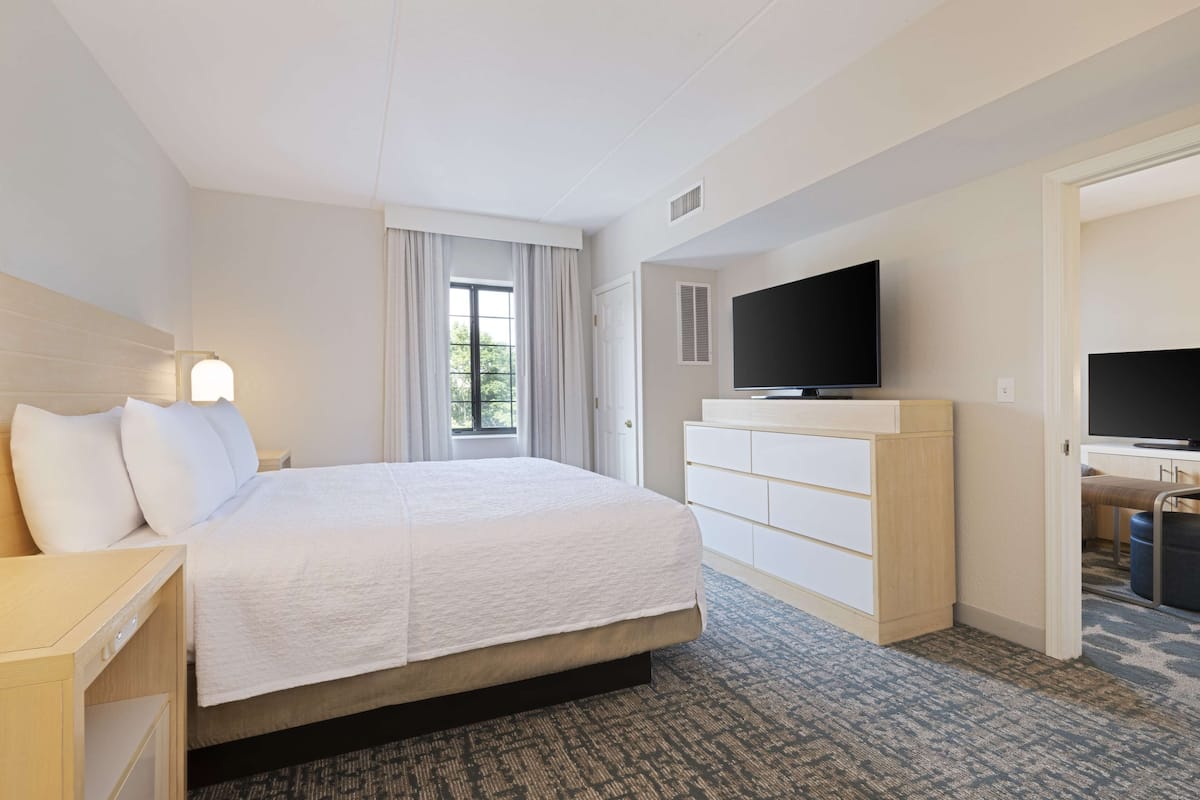 A hotel room with a large bed, a TV, a dresser, and a door leading to another room.