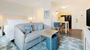 Moderni hotellihuone, jossa on sänky, sohva, pieni pöytä, roskakori, pieni keittiö ja televisio.