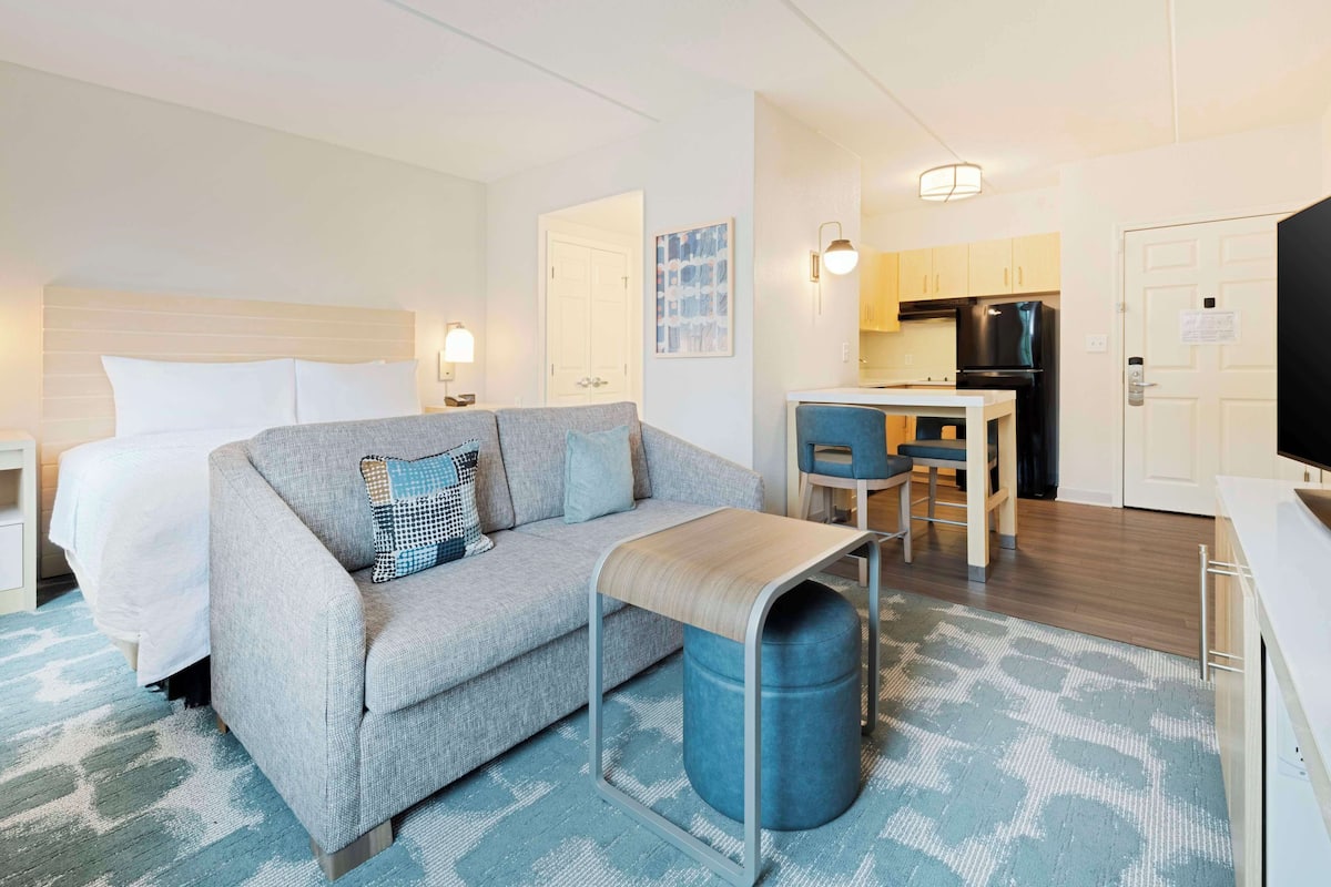 A modern hotel room with a bed, a sofa, a small table, a trash bin, a kitchenette, and a TV.