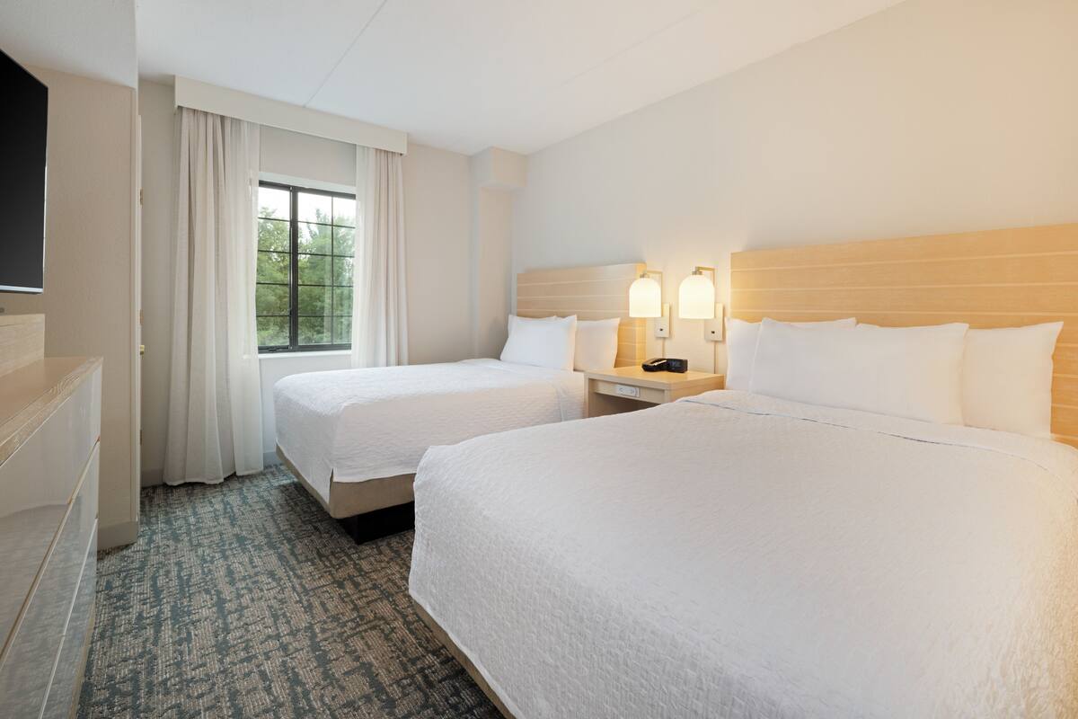 A hotel room with two beds, a TV, a window with curtains, and a bedside table with lamps.