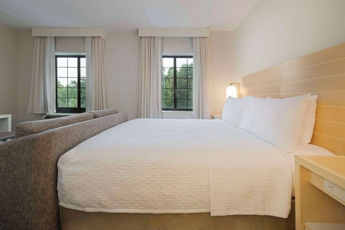 A hotel room with a large bed, a sofa, two windows with curtains, and a nightstand with a lamp.