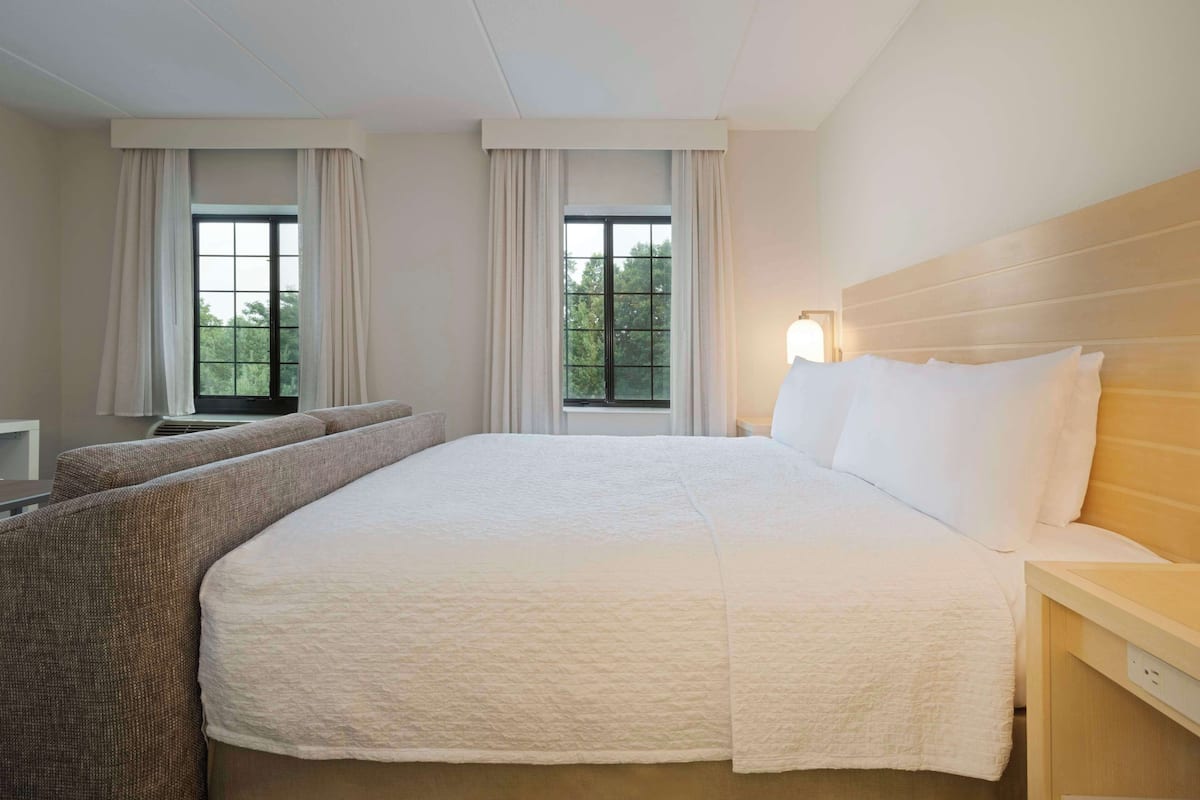 A hotel room with a large bed, a sofa, two windows with curtains, and a nightstand with a lamp.