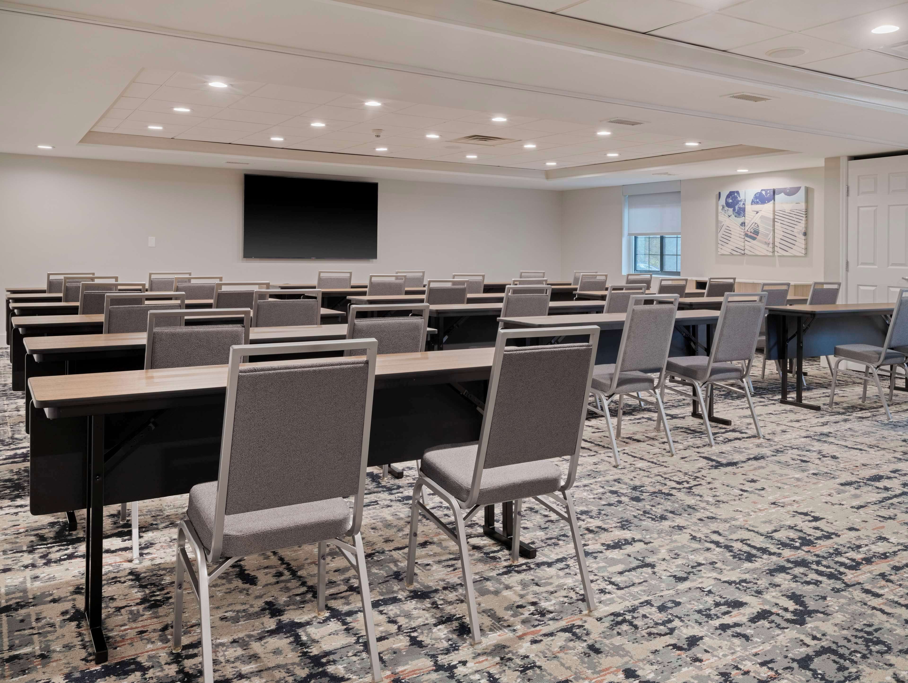 A conference room with rows of tables and chairs, a large screen, and a patterned carpet.