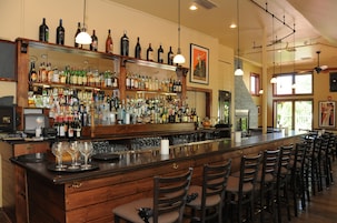 Una zona de bar con una barra alargada, varias estanterías llenas de botellas y varias sillas.
