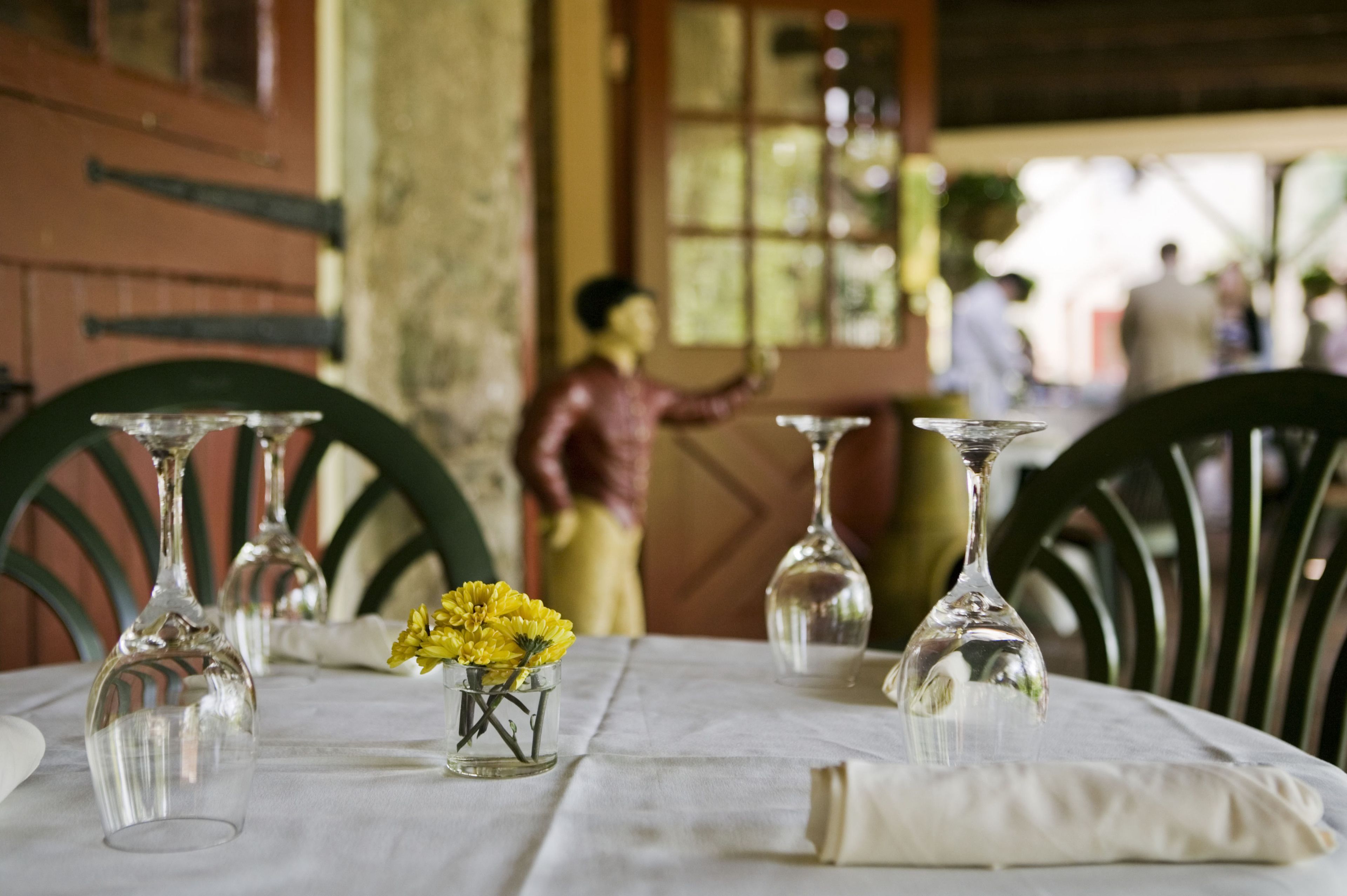A dining table with clear glassware and a small vase of yellow flowers, set in a room with wooden chairs and a rustic ambiance.