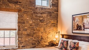 Une chambre avec un mur en pierre, un grand lit agrémenté d’oreillers à motifs, une fenêtre avec des stores et un tableau encadré accroché au mur.