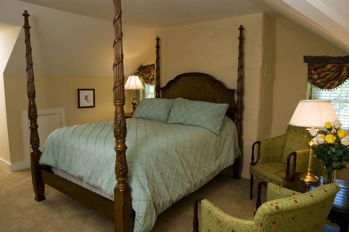 Ein Bett mit Baldachin, ausgestattet mit einer gesteppten Tagesdecke, einem Holzkopfende und einem Nachttisch mit Lampe und Blumen. Außerdem steht ein Sessel und ein Tisch mit einer Blumenvase dort.