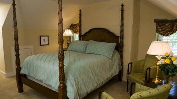 A four-poster bed with a quilted bedspread, a wooden headboard, and a nightstand with a lamp and flowers. There is a chair and a table with a vase of flowers.